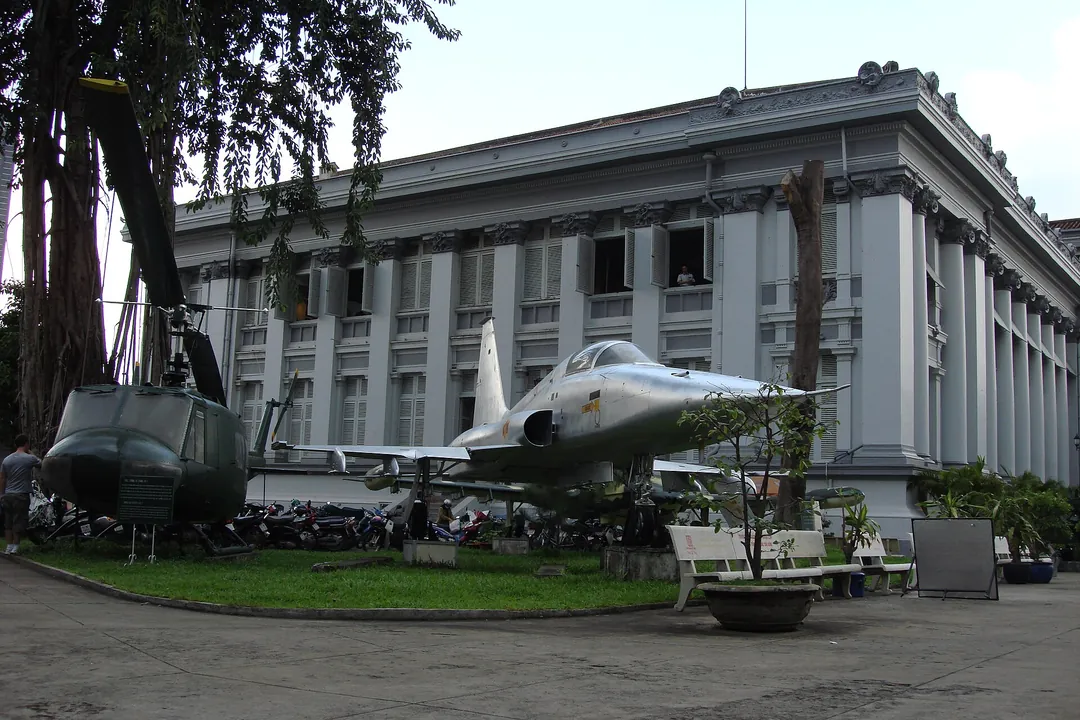 A Huey helicopter and a fighter jet parked on the lawn beside the grey museum building