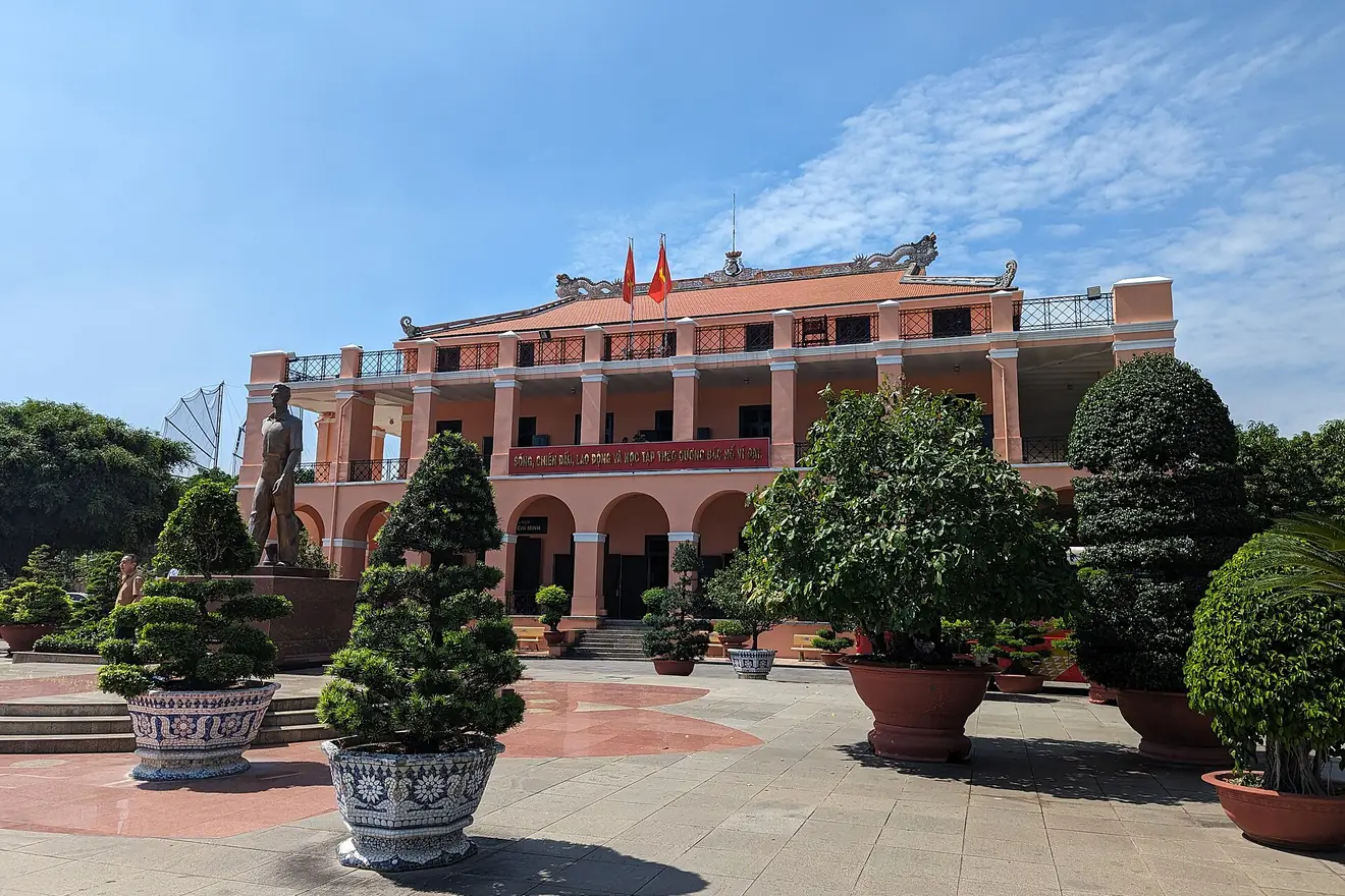 The pink Nhà Rồng building behind a plaza of potted trees and a bronze statue