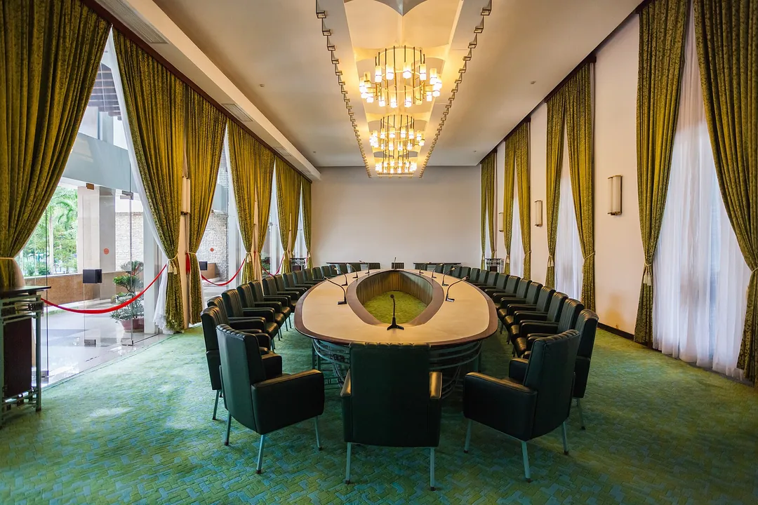 An oval table ringed by green leather chairs in a large room with gold curtains