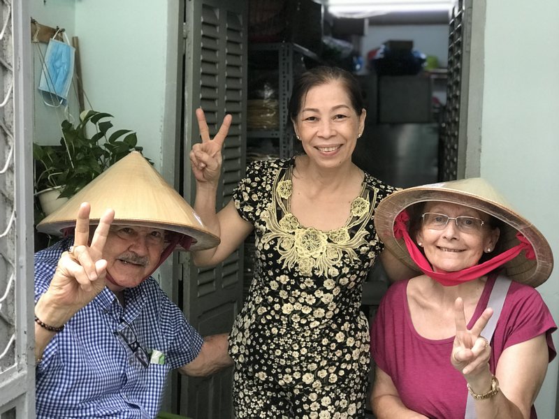 Two guests in conical nón lá hats with a local woman at her front door in a Saigon neighbourhood