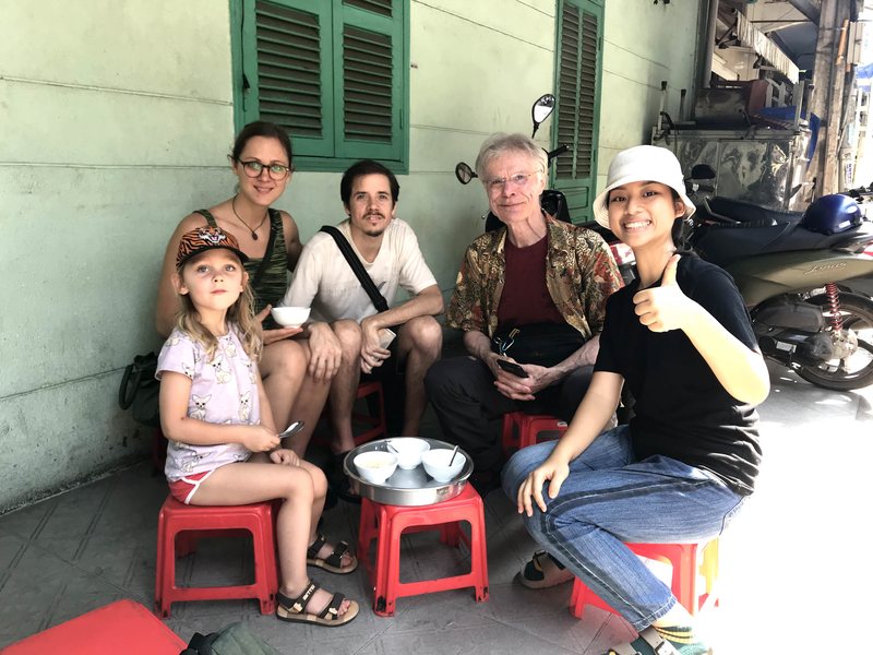 Our guide and a family on red plastic stools with bowls of a local snack, against green shutters in a Saigon neighbourhood