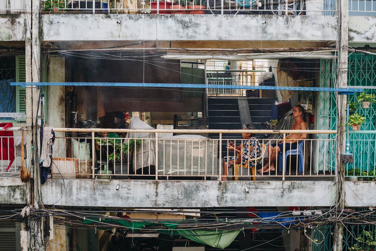 An older woman and man sitting on plastic chairs on a balcony of the old apartment block, its concrete walls stained grey and strung with wires