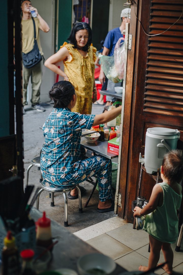 A woman in a pyjama set at a breakfast table in a shop doorway, a toddler watching from the step, neighbours in the alley behind