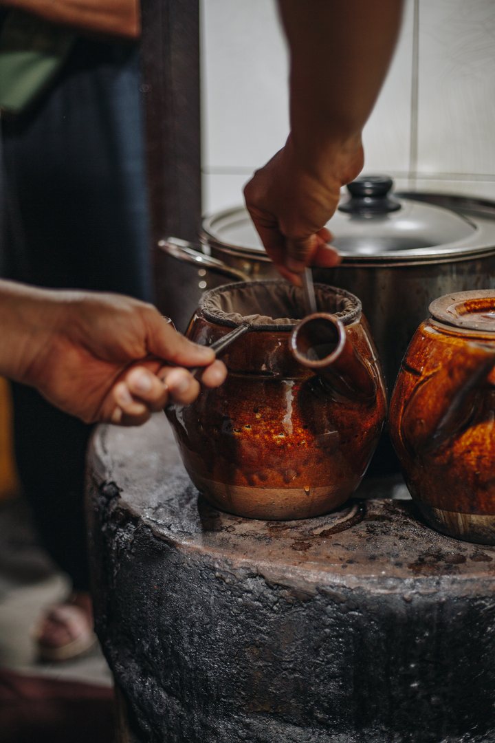 Hands working a cloth coffee filter over glazed clay pots on a soot-blackened charcoal stove