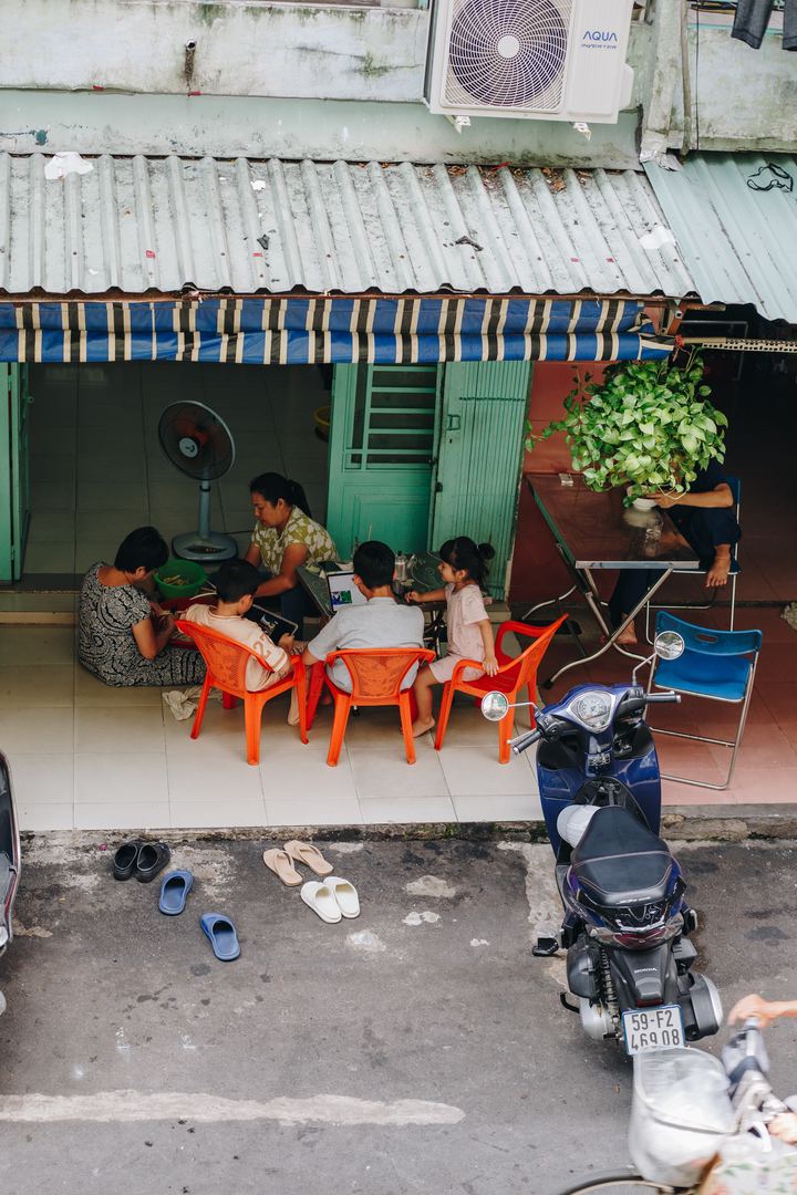 A family at a low table outside their ground-floor home in the apartment block, kids on a tablet and laptop, a woman sorting vegetables, seen from above