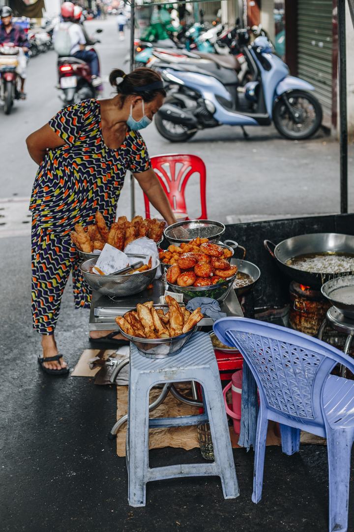 A vendor in a face mask at a street-side fritter stall: chuối chiên, sesame-glazed cakes and other fritters, scooters passing behind