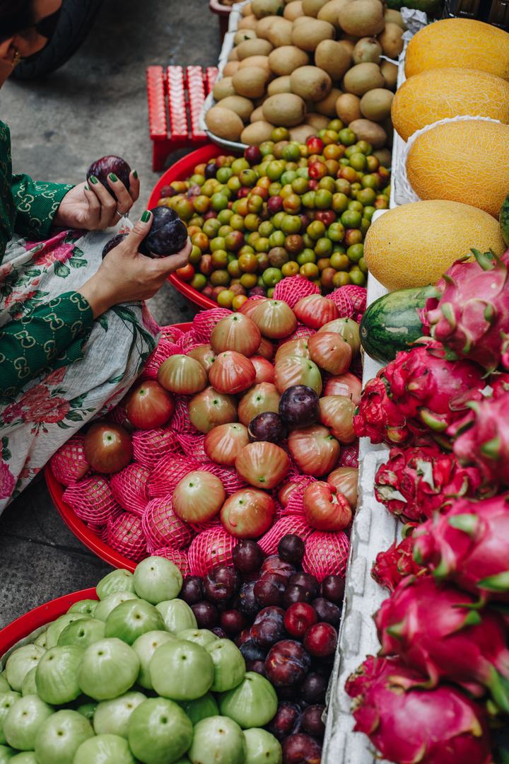 A woman holding up two plums at a fruit stall piled with melons, sapodilla, dragon fruit and small green and red fruit