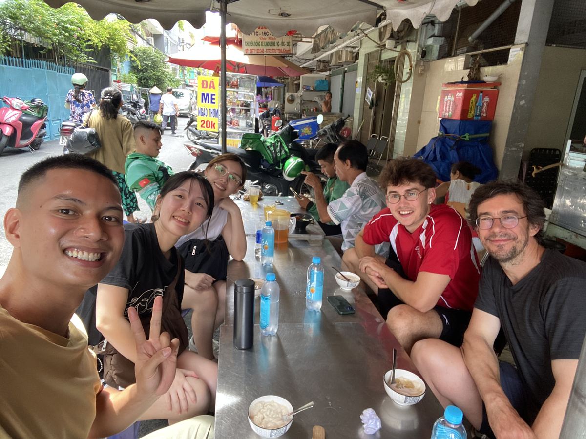Hieu taking a selfie with four guests over bowls of tàu hũ at a long steel table on the street, motorbikes parked behind