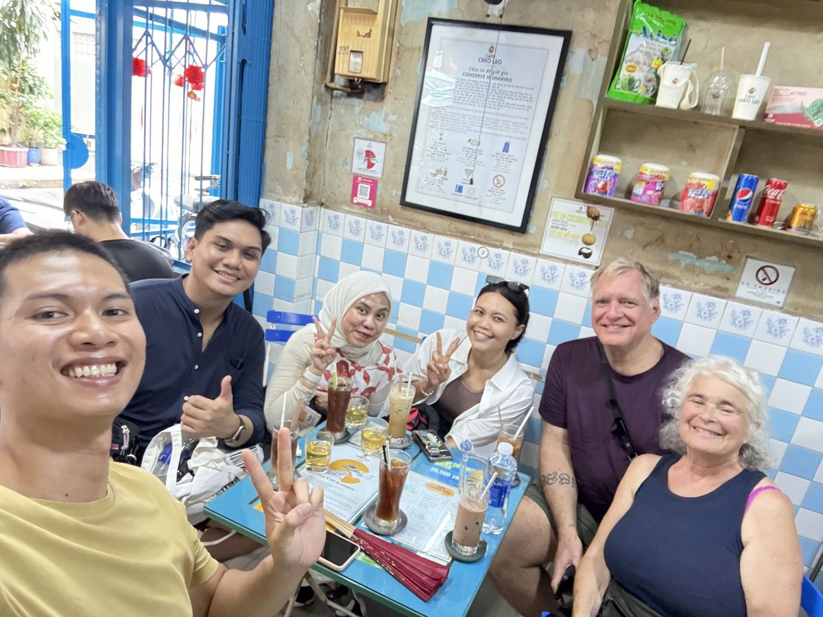 Hieu taking a selfie with five guests over iced coffees at Café Chéo Leo, blue and white tiles on the wall behind