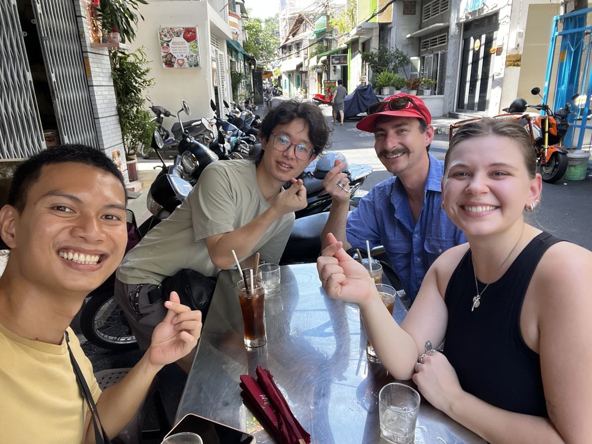 Hieu taking a selfie with three guests at a steel table in a sunny alley, iced drinks in front of them