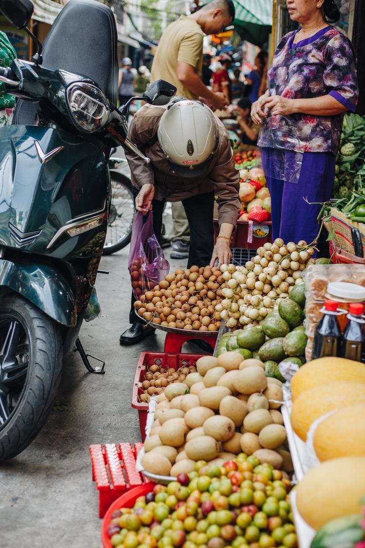 A woman in a motorbike helmet picking longan into a bag at a street fruit stall while the vendor looks on