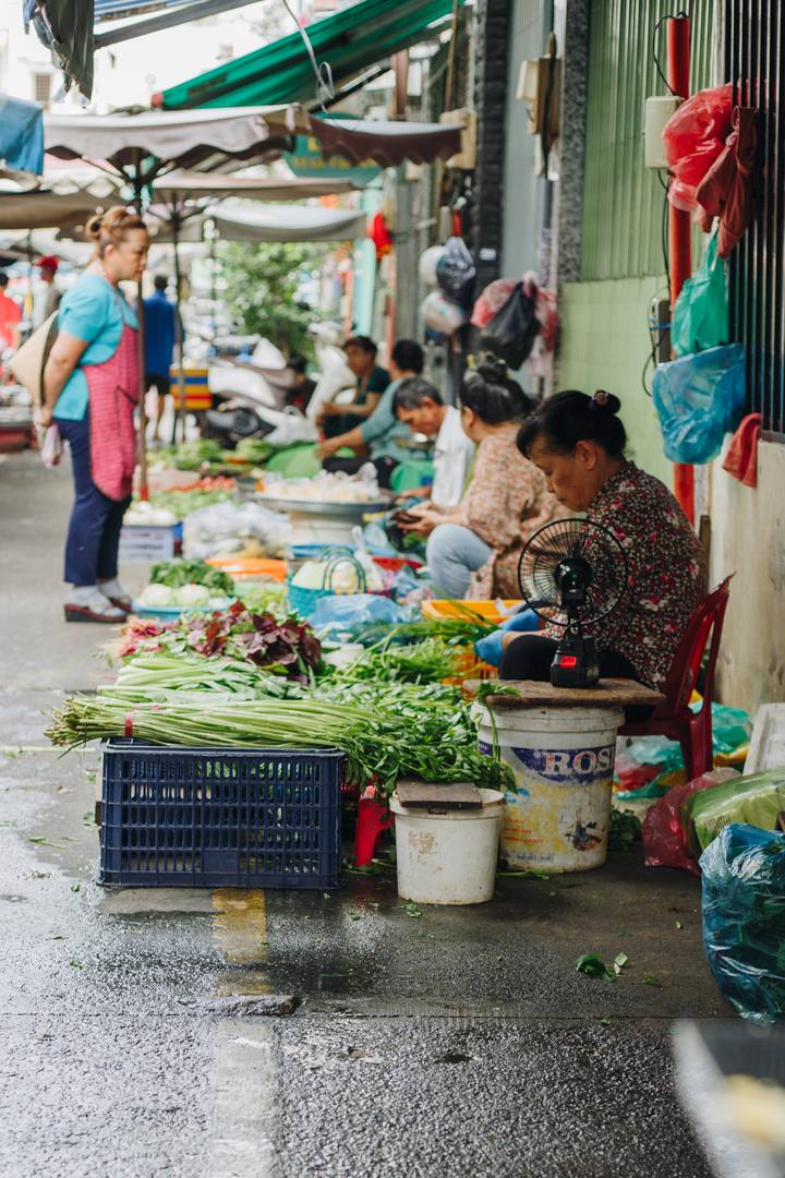 Vendors on low stools beside bundles of water spinach and red amaranth on a wet market lane