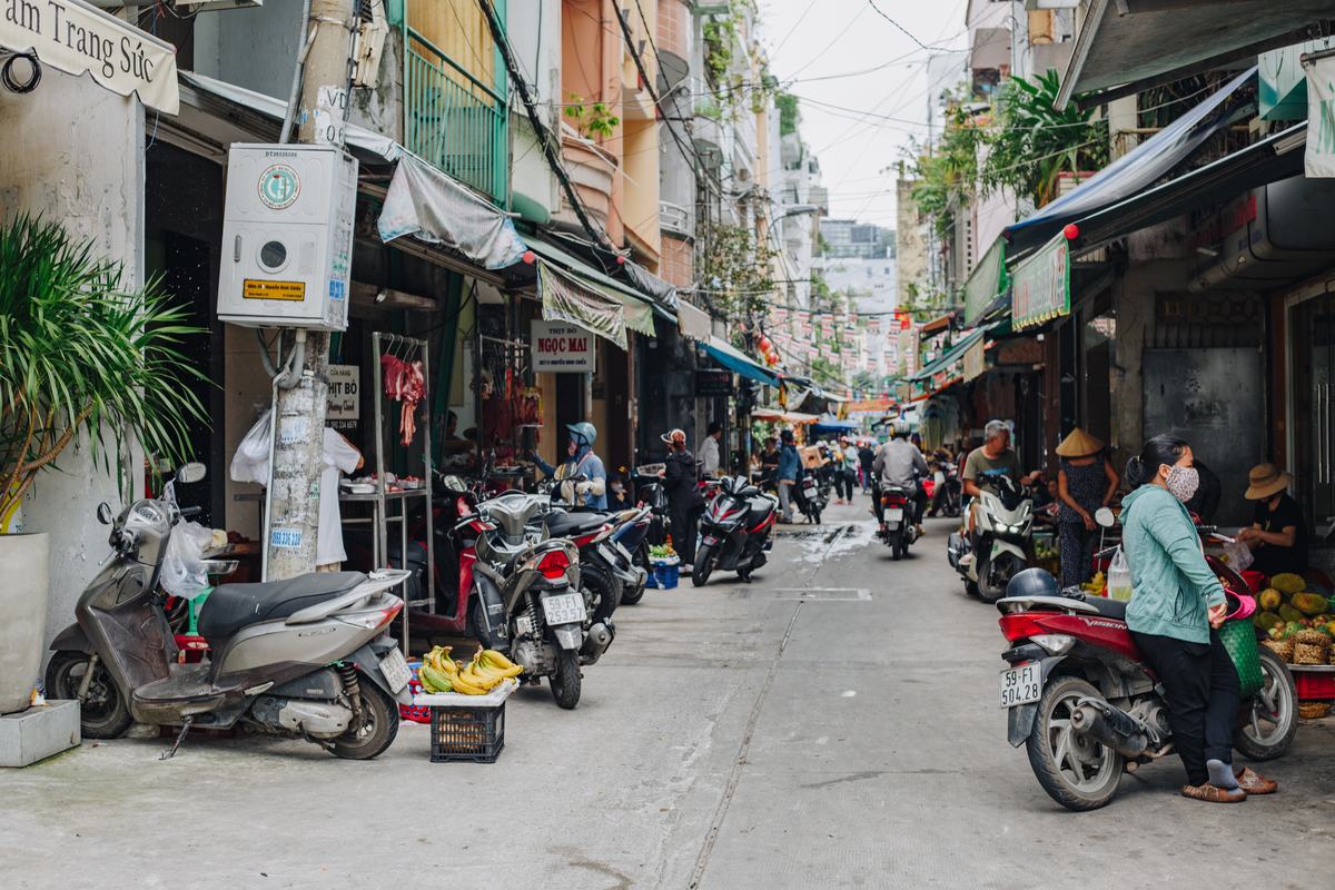 A market street in the morning: scooters parked outside a beef shop, meat hanging at the stalls, people riding through under strings of small flags