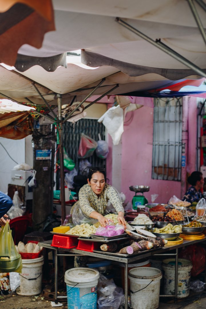 A vendor reaching across a stall of taro, cassava and bowls of prepared ingredients under the market umbrellas, a pink wall behind