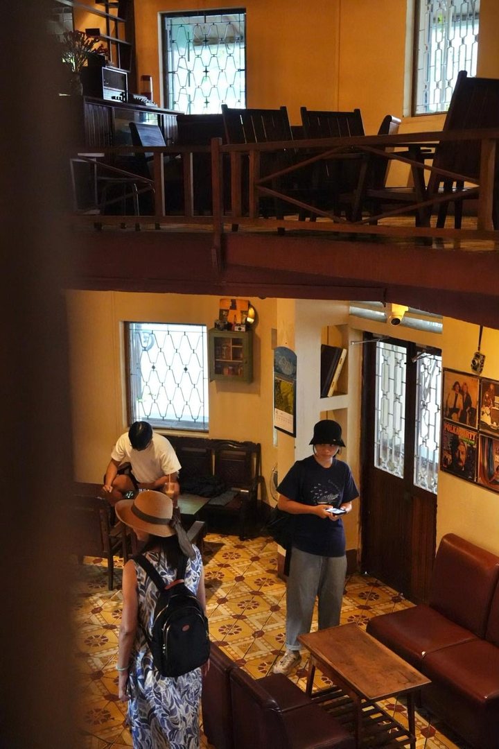 Spring and a guest in an old café, seen from the wooden mezzanine above: patterned tile floor, leather sofas and framed photos on the wall