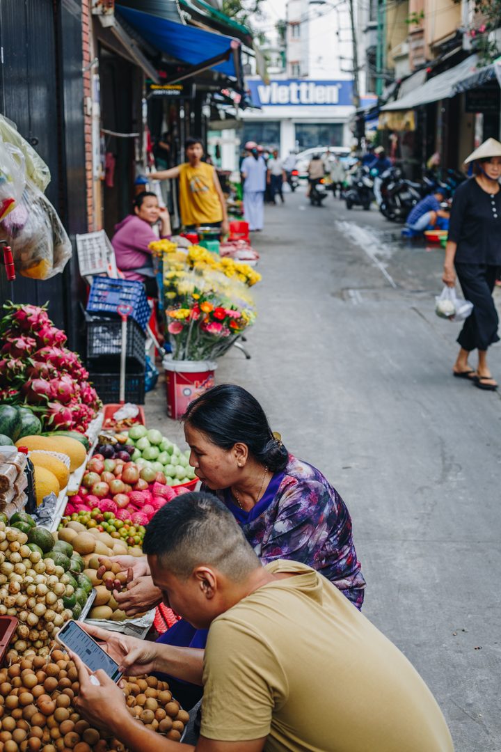 One of our team crouched at a fruit stall with a phone in hand while the vendor sorts longan, flower sellers and the market street behind