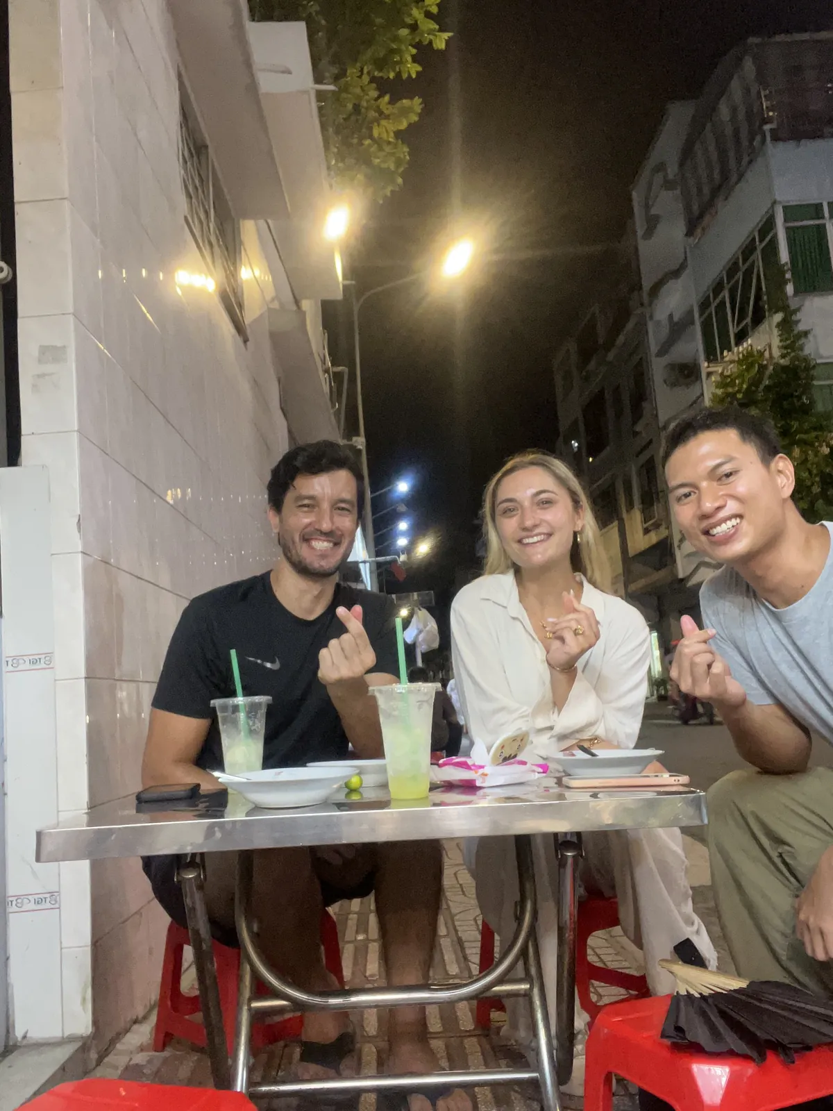 Hieu and two guests at a steel street table at night, with plates, iced drinks and red plastic stools