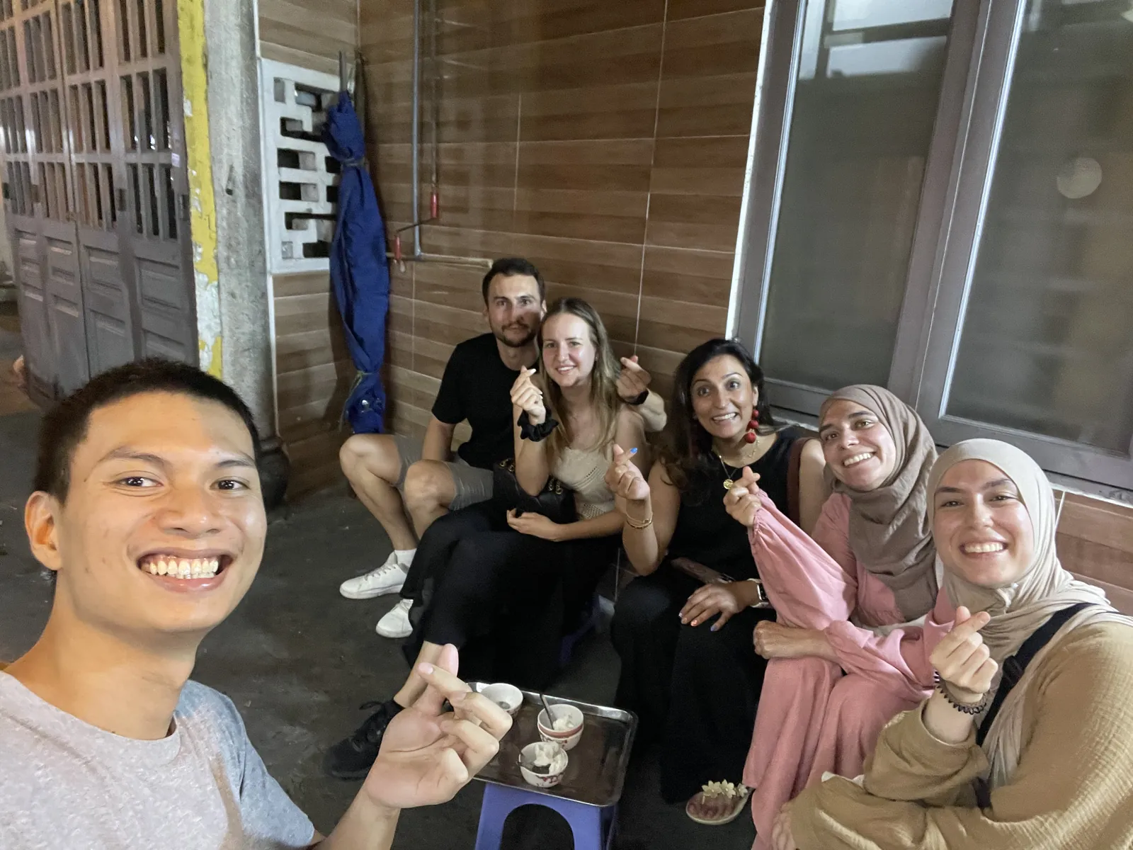 Hieu taking a selfie at night with five guests on low plastic stools by the street, small bowls on a tray in front of them