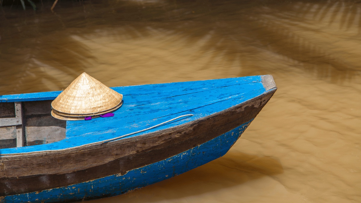A wooden sampan boat with a conical hat resting on the bow, on the brown water of the Mekong Delta