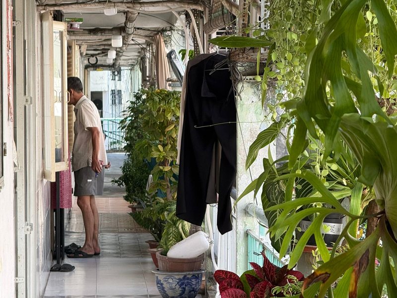 A resident at his door in a bright apartment corridor lined with potted plants and shoes
