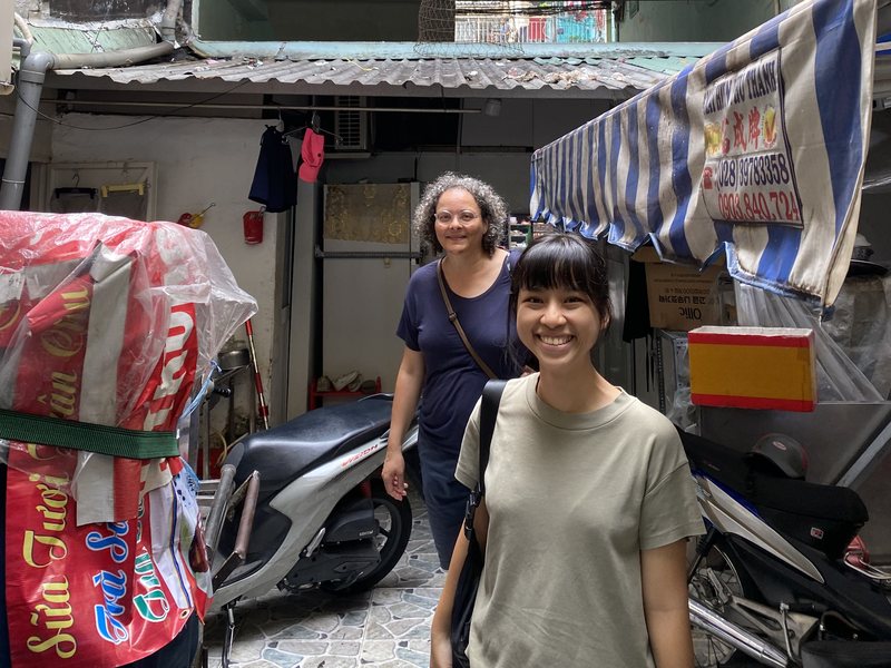 Two women smiling at the foot of an old apartment block, parked motorbikes and a striped awning beside them