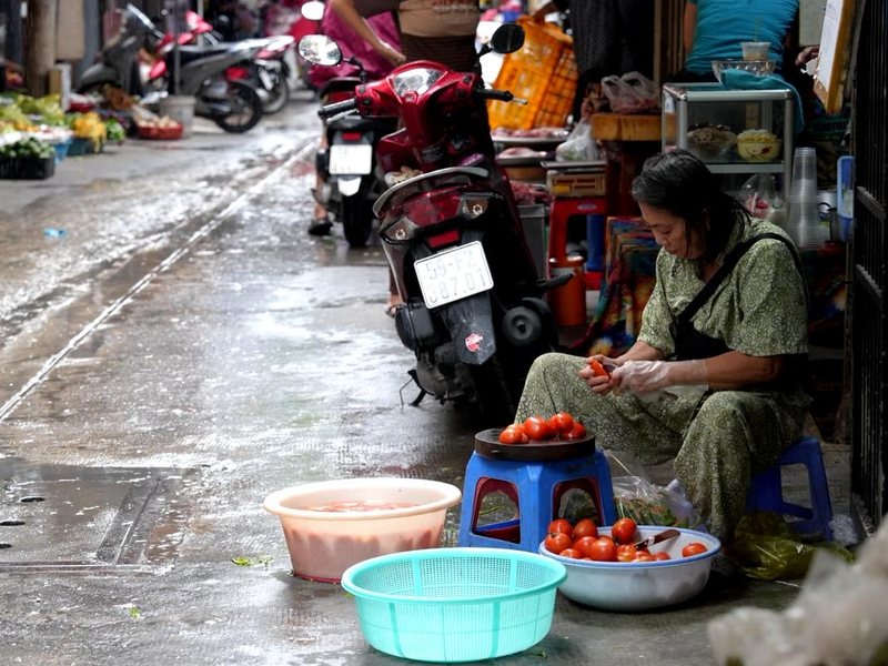 A vendor sorting tomatoes on a low stool in a wet market lane, motorbikes parked along the alley