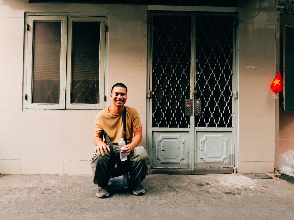 One of our team sitting on a low stool outside a house with a white metal door in a quiet Saigon street, smiling