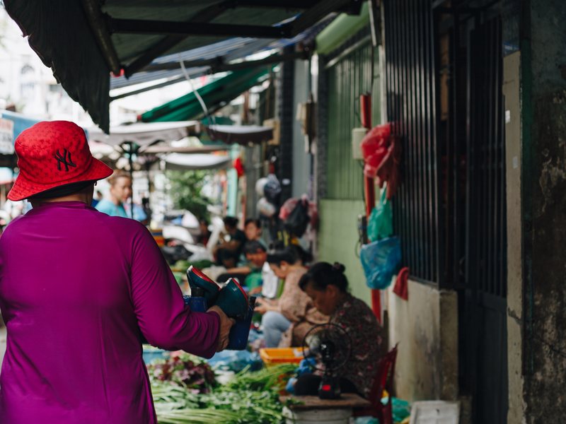 A shopper in a red hat walking a narrow market lane past vendors selling greens