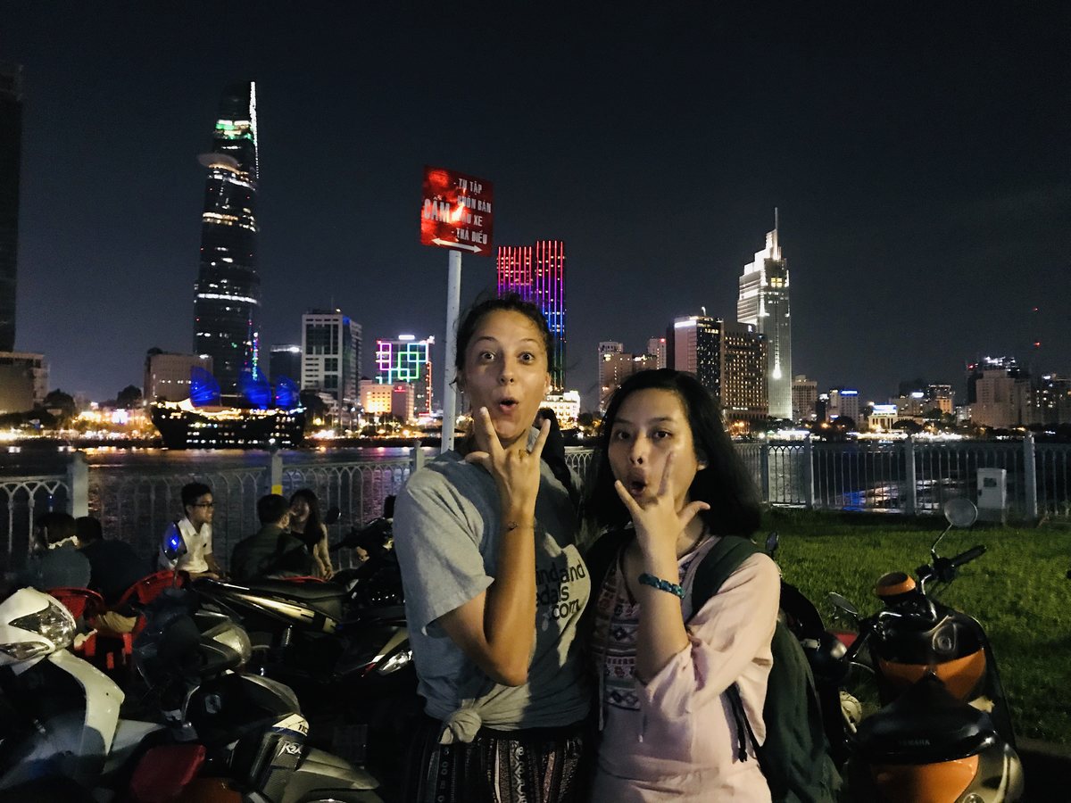 Two women posing by the Saigon River at night, Bitexco Tower and the lit-up skyline behind them
