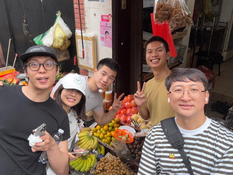 Hieu with four guests at a fruit stall piled with bananas, oranges and longan