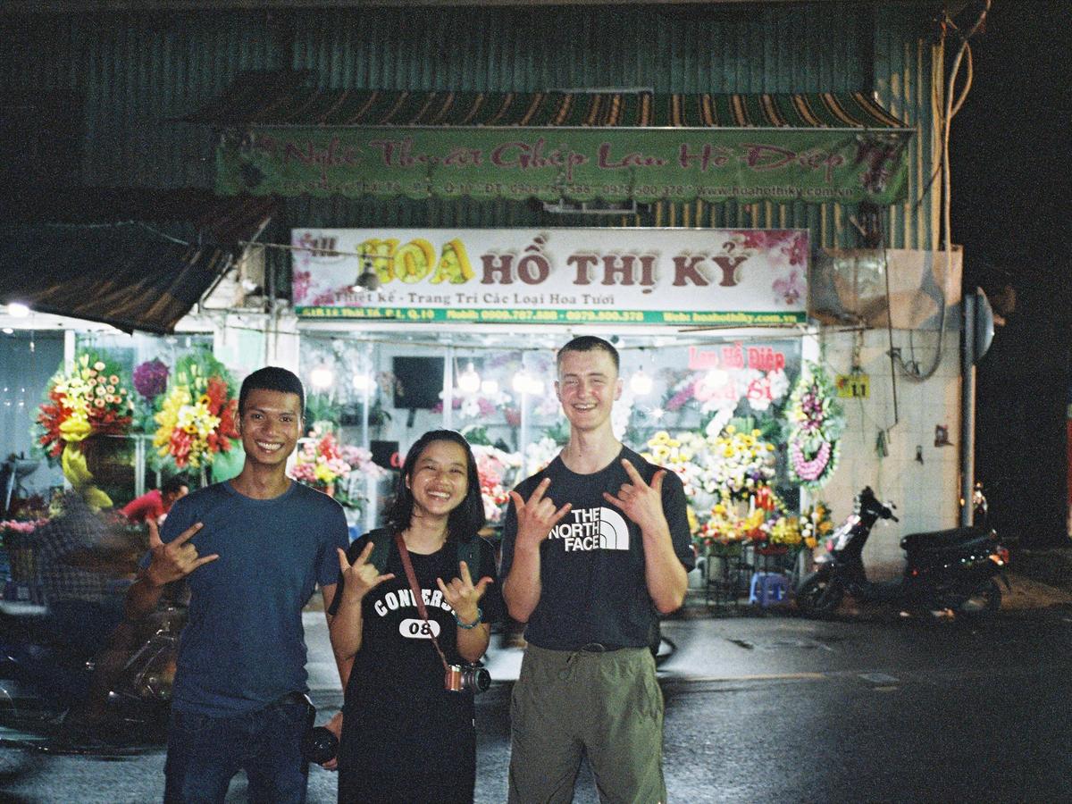 Three people at night outside a flower stall at Ho Thi Ky flower market, the stall's sign lit up behind them