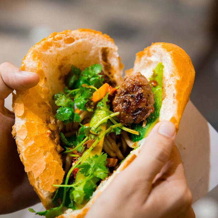 A bánh mì torn open in two hands: a grilled meatball, noodles, pickled carrot, cucumber and coriander inside