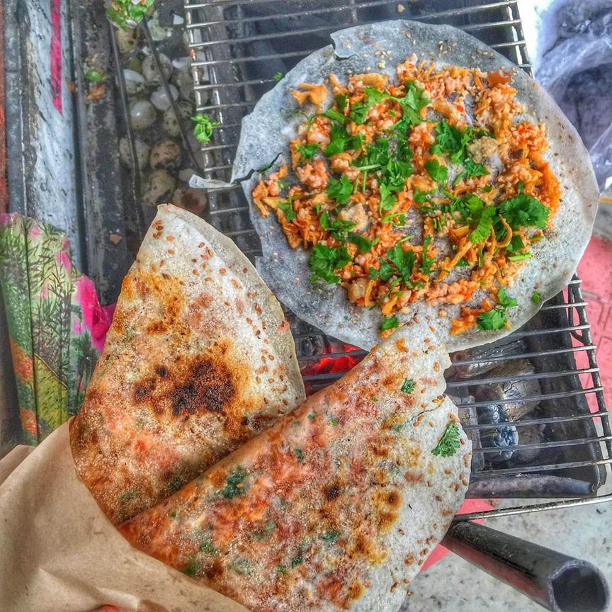Bánh tráng nướng on a charcoal grill: one sheet still cooking under chilli toppings and coriander, two finished ones folded in half in a paper wrap, crusted with sesame
