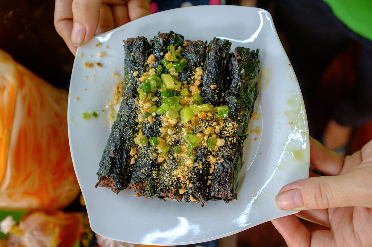A plate of six bò lá lốt rolls held in both hands, the betel leaves grilled almost black, topped with crushed peanuts and spring onion oil