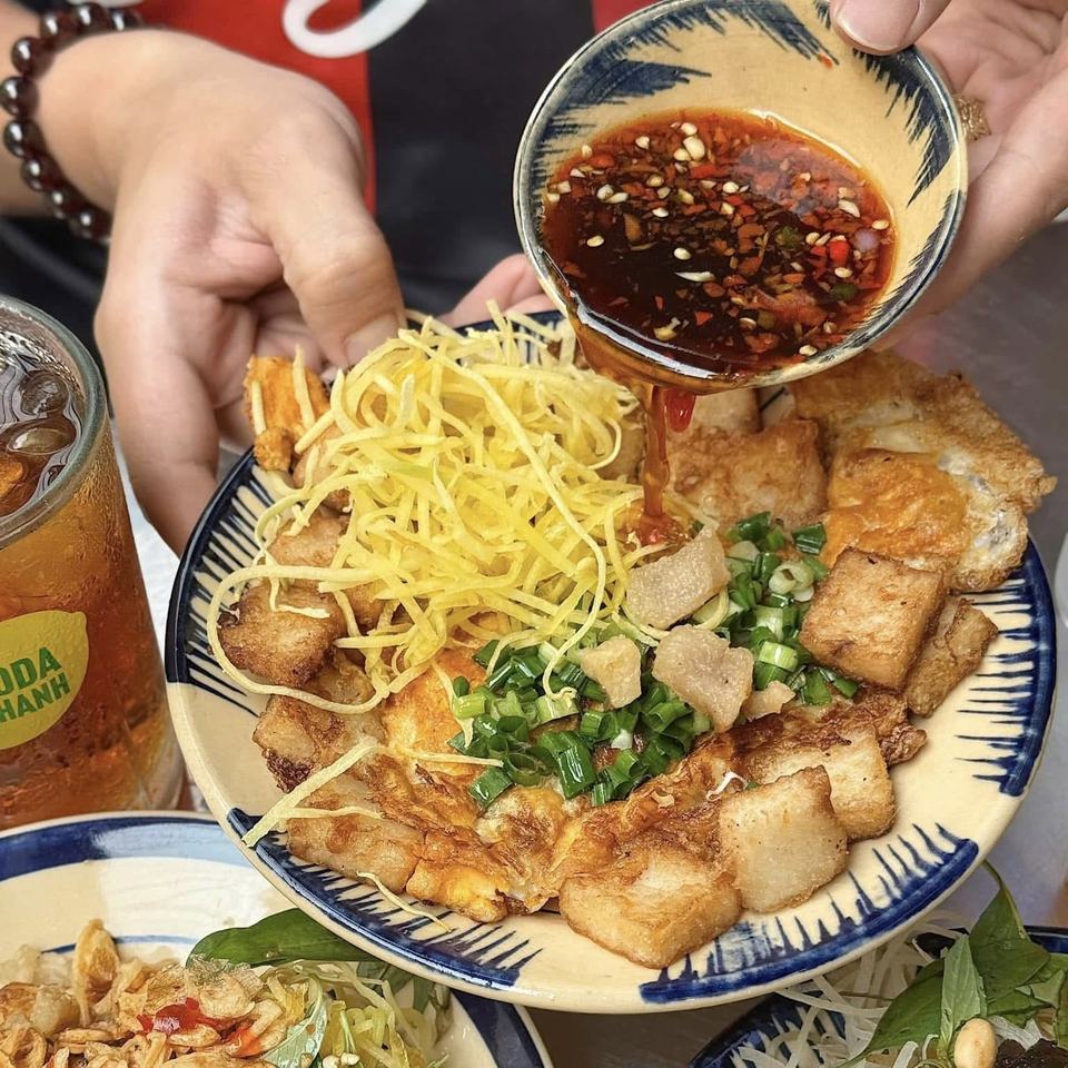 Bột chiên on a blue-rimmed plate: fried rice cake cubes and egg under shredded papaya and spring onion, with the soy and chilli dip being poured over