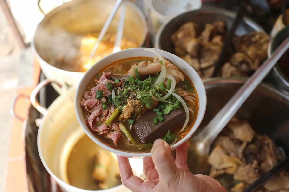 A bowl of bún bò Huế held up over the stall's pots: rare beef slices, pork hock, a cube of pork blood, onion and herbs in a red chilli broth