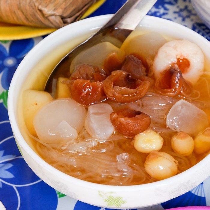 A bowl of chè with dried longan, lychee, lotus seeds and jelly in syrup, a spoon resting in it