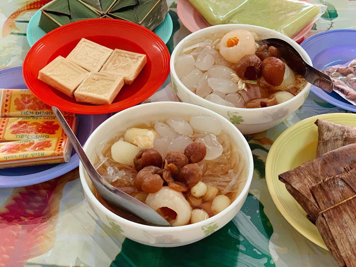 Two bowls of chè with dried longan, lychee, lotus seeds and jelly over ice, next to mung bean cakes and leaf-wrapped rice cakes
