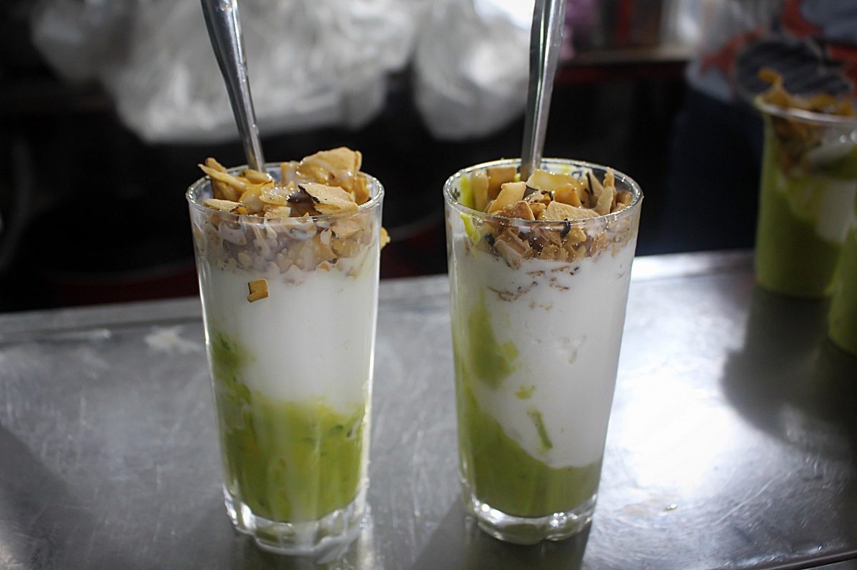 Two glasses of kem bơ on a steel counter: mashed avocado at the bottom, a layer of white ice cream, toasted coconut flakes on top